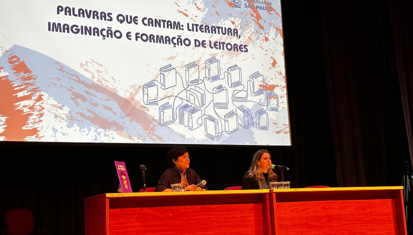 Palco com duas pessoas em uma mesa de madeira e um telão branco de fundo