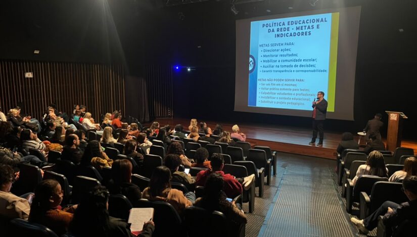 Homem em pé no canto direito do palco de um teatro, segurando um microfone e realizando uma palestra. À sua frente, diretores estão sentados em cadeiras, acompanhando atentamente a apresentação em um ambiente de reunião e alinhamento institucional.