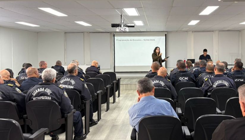Fotografia mostra uma mulher a frente de um auditório. Ela fala com as pessoas da platéia com um microfone. A maiorria das pessoas que estão sentadas estão com o uniforme da Guarda Civil Metropolitana (GCM).