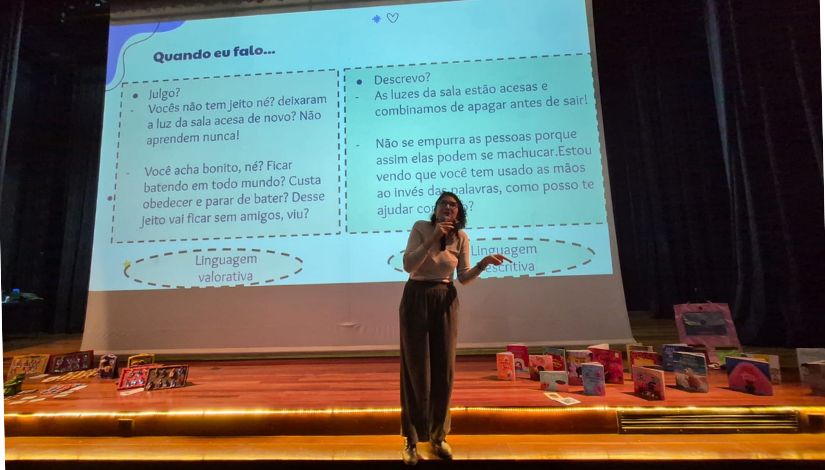 Fotografia de uma mulher ao palco falando no microfone. Ao fundo, telão com conteúdo projeto.