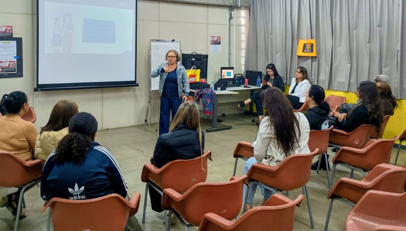 Em uma sala de formação, uma mulher em pé apresenta uma atividade para um grupo de participantes sentados em cadeiras organizadas em semicírculo. Ao fundo, há uma tela de projeção, um computador e uma mesa de apoio. Os participantes observam atentamente a apresentação, em um ambiente interno com cortinas claras e paredes neutras.