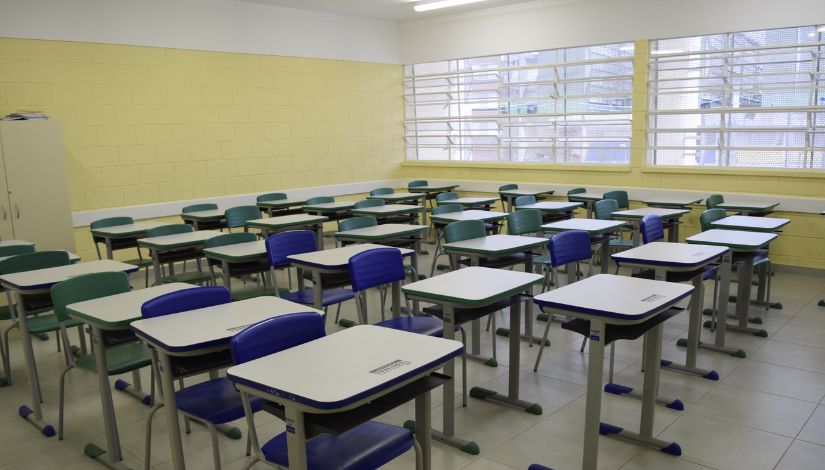 otografia mostra uma sala de aula vazia, com carteiras e cadeiras organizadas em fileiras. O ambiente é amplo, iluminado por grandes janelas laterais protegidas por grades, possui paredes em tom amarelo-claro, piso cinza e um armário branco ao fundo. Não há pessoas na imagem.