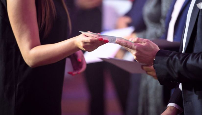 Pessoa entrega um certificado a outra durante uma cerimônia de premiação. A imagem mostra apenas as mãos dos participantes e o momento da entrega do documento, simbolizando o reconhecimento de profissionais da educação.