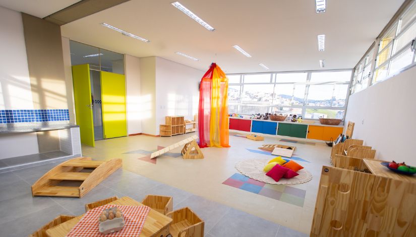 Fotografia de uma sala de Centro de Educação Infantil com móveis pequenos de madeiras para bebês e uma tenda de tule laranaja e vermelho ao centro.