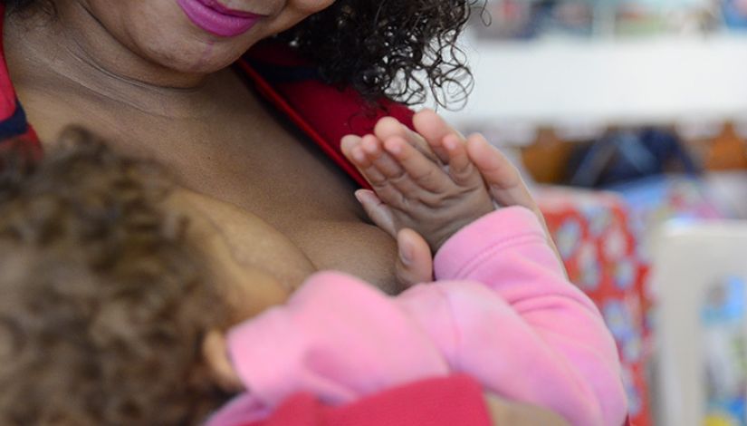 Em primeiro plano, uma mulher amamenta um bebê, que repousa em seus braços com uma das mãos apoiada sobre seu peito. A cena representa um momento de cuidado, acolhimento e fortalecimento do vínculo entre mãe e criança, tema da campanha Agosto Dourado.