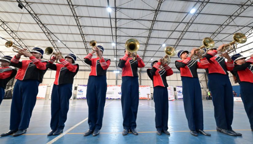 Estudantes integrantes de uma banda marcial se apresentam em quadra coberta, posicionados em formação e tocando instrumentos de metal, como trompetes e trombones. Os participantes vestem uniformes nas cores vermelha, azul-marinho e preta, com chapéus brancos. Ao fundo, vê-se o espaço amplo do ginásio, com iluminação no teto e banners do evento.