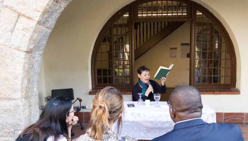 Mulher sentada atrás da mesa lendo um livro de capa ver que segura com a mão esquerda.