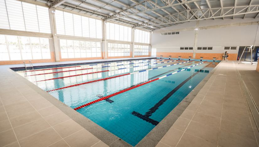Fotografia mostra a piscina coberta do CEU Silvio Santos. O espaço é amplo, iluminado por grandes janelas laterais que permitem a entrada de luz natural. A piscina possui raias delimitadas por flutuadores nas cores vermelha, branca e azul, além de marcações escuras no fundo que orientam a prática da natação. Ao redor da piscina há piso antiderrapante, escadas de acesso à água, plataformas de saída e equipamentos de apoio aos treinamentos. A imagem destaca a infraestrutura esportiva destinada às atividades de natação e promoção do esporte nos Centros Educacionais Unificados (CEUs).