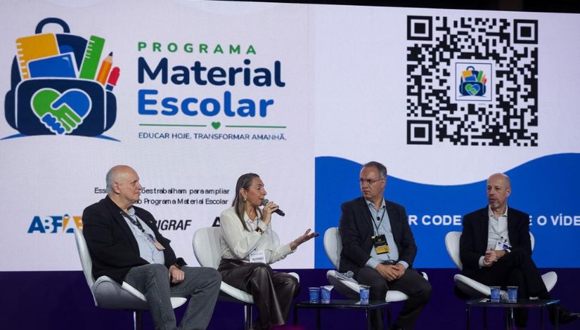 Quatro participantes estão sentados em poltronas durante um painel em um evento, enquanto uma mulher fala ao microfone e gesticula. Ao fundo, um telão exibe a identidade visual do Programa Material Escolar, com logotipo, QR Code e elementos gráficos relacionados à iniciativa.