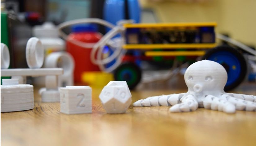 Em primeiro plano, objetos produzidos em impressora 3D, como um polvo, um dado geométrico e peças didáticas, estão dispostos sobre uma mesa. Ao fundo, equipamentos e protótipos de robótica aparecem desfocados, representando atividades voltadas à inovação, tecnologia e aprendizagem prática em ambiente educacional.