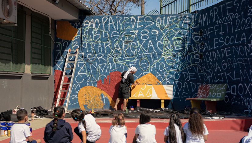 Fotografia mostra um artista pintando um mural em grande escala no muro de uma escola da Rede Municipal de Ensino. A obra, em processo de criação, apresenta desenhos, letras e formas geométricas sobre um fundo azul, com áreas em tons de amarelo e laranja. O artista trabalha em frente ao mural utilizando materiais de pintura, enquanto uma escada está apoiada ao lado da parede. Em primeiro plano, estudantes, vistos de costas e sentados no pátio, acompanham a atividade artística. A cena representa a integração entre arte, educação e participação da comunidade escolar