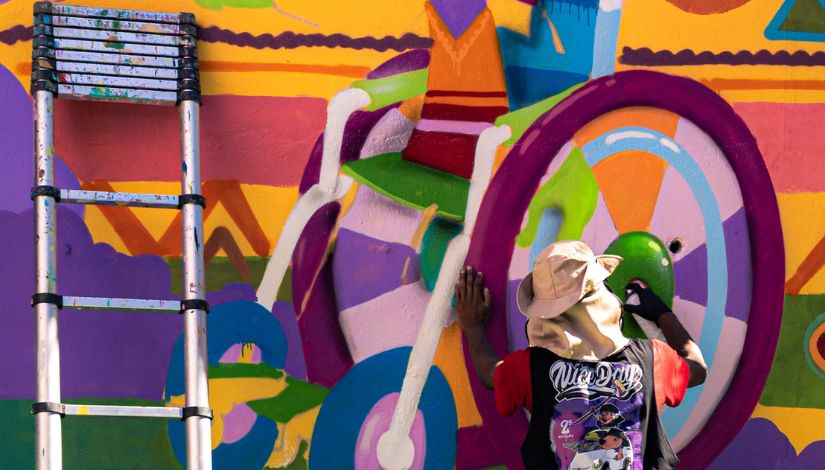 Fotografia mostra um artista urbano pintando um mural colorido no muro de uma escola da Rede Municipal de Ensino. Visto de costas, o artista utiliza tinta spray para dar forma à obra, composta por figuras abstratas e elementos geométricos em tons vibrantes de roxo, laranja, verde, azul, rosa e amarelo. À esquerda da imagem, uma escada está apoiada junto ao mural, indicando o processo de criação artística. A cena destaca a valorização da arte urbana como instrumento de expressão, transformação dos espaços escolares e fortalecimento das ações culturais na educação.