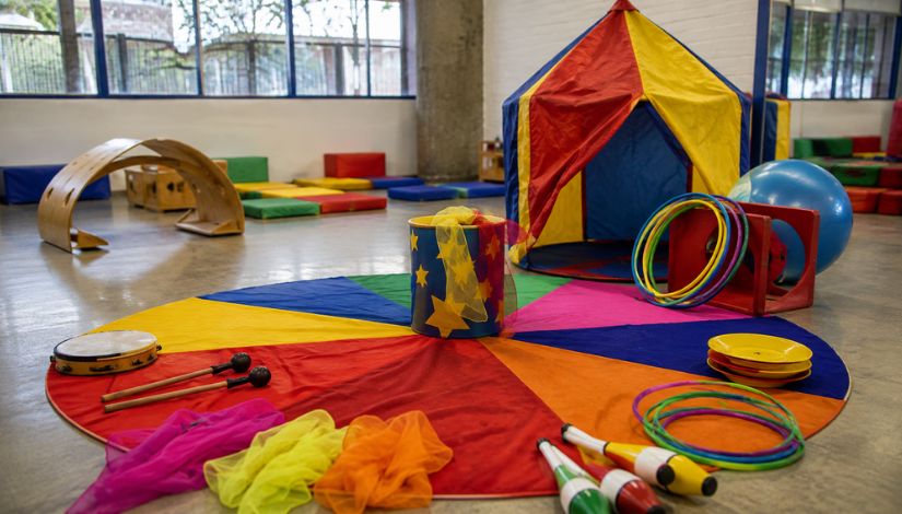 Fotografia ilustrativa de um espaço preparado para atividades culturais voltadas à primeira infância. No centro do ambiente há um tapete circular colorido, cercado por instrumentos de musicalização, como tambor e baquetas, além de materiais circenses, incluindo claves de malabarismo, bambolês, pratos de equilíbrio, tecidos coloridos e um balde decorado com estrelas. Ao fundo, destaca-se uma pequena tenda de circo nas cores vermelha, amarela e azul, além de colchonetes, estruturas de psicomotricidade e brinquedos distribuídos pelo espaço. A sala é ampla, iluminada por grandes janelas, e representa um ambiente lúdico destinado a experiências de teatro, musicalização, circo e desenvolvimento infantil.