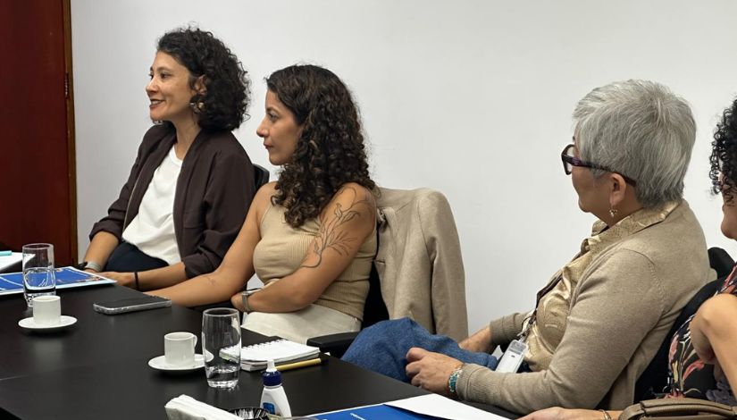 Três mulheres aparecem sentadas em cadeira na frente de uma mesa marrom
