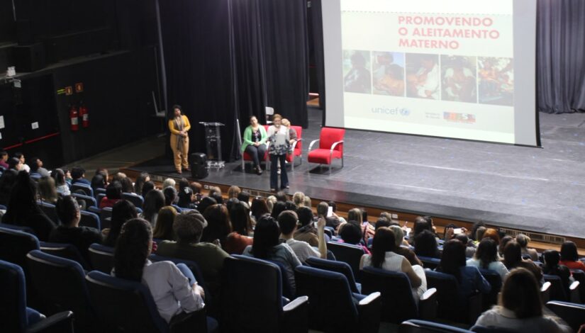 Auditório com público acompanhando uma apresentação sobre aleitamento materno. No palco, três mulheres participam da atividade e, ao fundo, um telão apresenta a mensagem “Promovendo o Aleitamento Materno”.