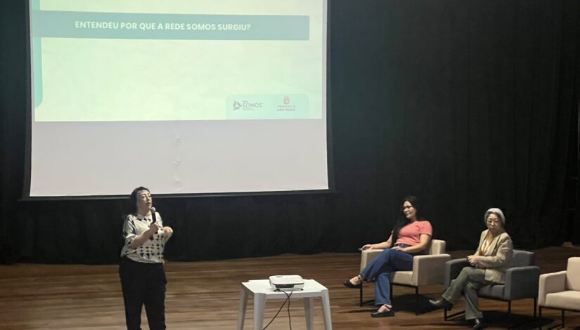 Em um auditório do CEU Inácio Monteiro, uma mulher fala ao microfone enquanto outras duas mulheres estão sentadas em poltronas no palco. Ao fundo, um telão exibe uma apresentação com o título “Entendeu por que a Rede Somos surgiu?”, além dos logotipos da Rede Somos e da Prefeitura de São Paulo.