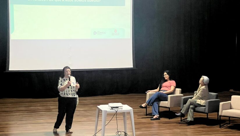 Em um auditório do CEU Inácio Monteiro, uma mulher fala ao microfone enquanto outras duas mulheres estão sentadas em poltronas no palco. Ao fundo, um telão exibe uma apresentação com o título “Entendeu por que a Rede Somos surgiu?”, além dos logotipos da Rede Somos e da Prefeitura de São Paulo.