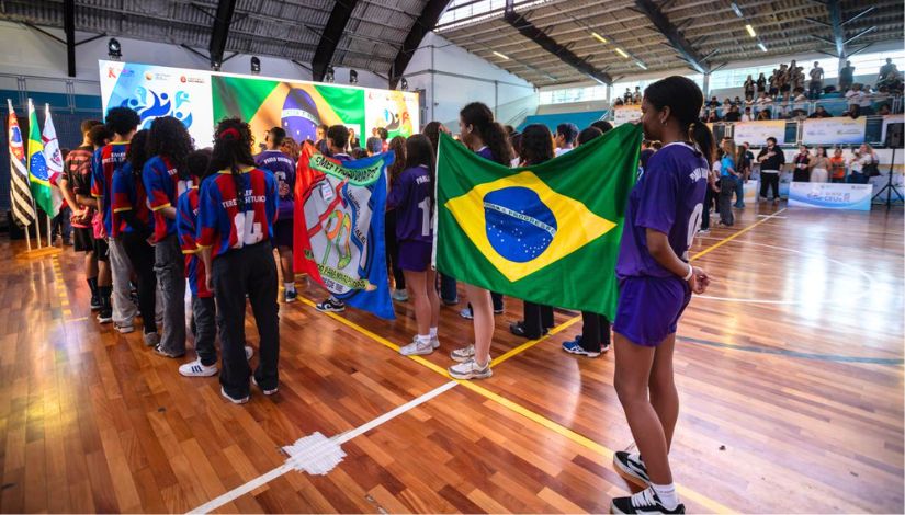 Estudantes seguram bandeiras durante cerimônia esportiva em ginásio.