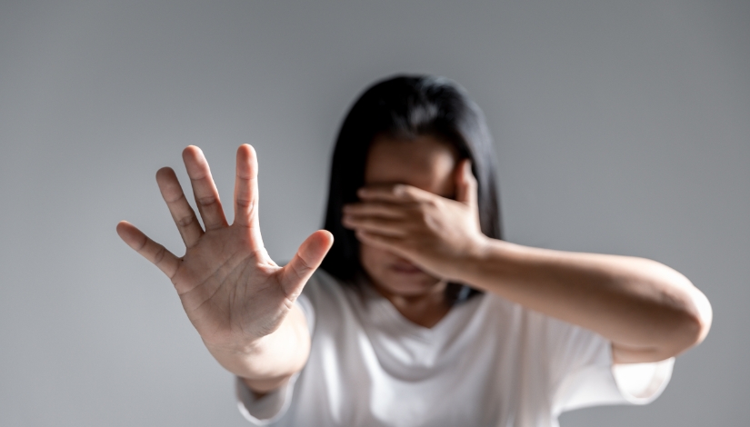 Mulher em primeiro plano estende uma das mãos em direção à câmera, enquanto cobre o rosto com a outra mão. Ao fundo, há uma parede em tom cinza. A imagem representa uma situação de pedido de ajuda ou proteção diante da violência.