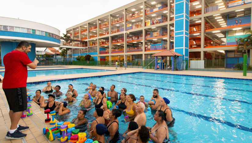Fotografia de uma piscina em um centro educacional. Na área da piscina, um grupo de adultos está dentro da água, participando de uma atividade orientada por um profissional que está de pé na borda. Ao redor, há materiais coloridos utilizados na atividade. Ao fundo, é possível ver o complexo educacional, com vários andares, áreas abertas e uma segunda piscina.