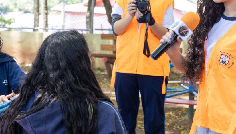 Fotografia de estudantes da Imprensa Jovem realizando uma entrevista ao ar livre. Em primeiro plano, uma estudante segura um microfone próximo a outra estudante, que está de costas. Ao fundo, outro estudante registra a atividade com uma câmera fotográfica. Os jovens usam coletes laranja e azul, identificados como integrantes da Imprensa Jovem.