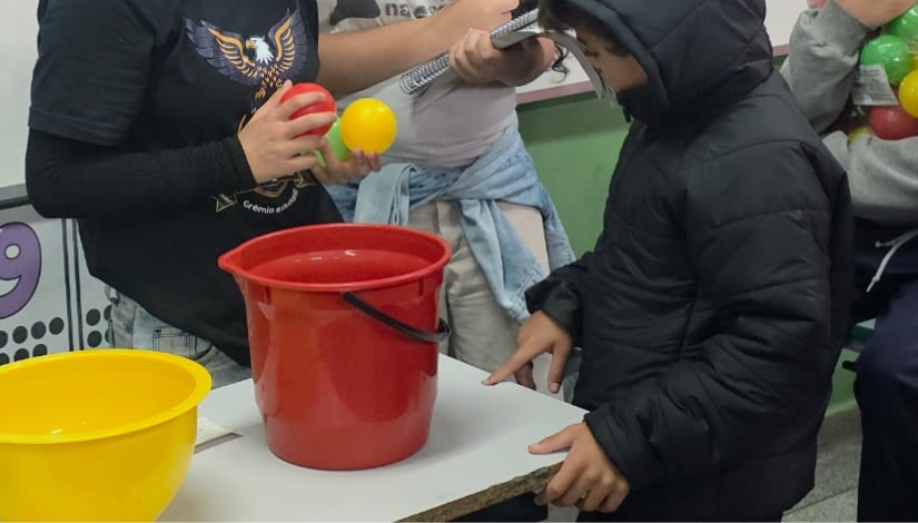 Fotografia mostra estudantes reunidos em torno de uma mesa durante uma atividade escolar. Um estudante, à direita, veste casaco preto e participa da atividade, enquanto outros estudantes, ao centro, seguram bolas coloridas, nas cores vermelha e amarela. Sobre a mesa há dois baldes, um vermelho e outro amarelo, utilizados na atividade. Ao fundo, aparecem materiais e elementos coloridos do ambiente escolar.