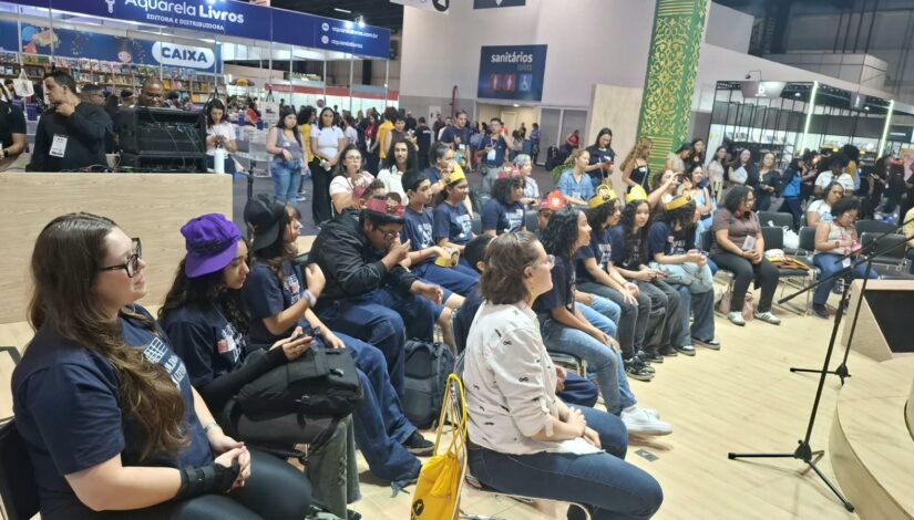 Público acompanha uma atividade durante a Bienal do Livro, em espaço interno do evento. Pessoas estão sentadas diante de um palco, enquanto outras acompanham a programação ao fundo, próximo aos estandes de livros.