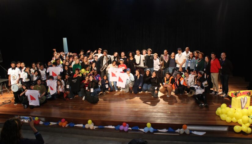 Fotografia de um grupo de estudantes, educadores e convidados reunidos no palco de um teatro, posando para uma foto coletiva. Algumas pessoas seguram camisetas e instrumentos musicais. Ao fundo, há cortinas pretas e iluminação de palco. Na parte inferior da imagem, aparecem balões coloridos e uma pessoa fotografando o grupo na plateia.