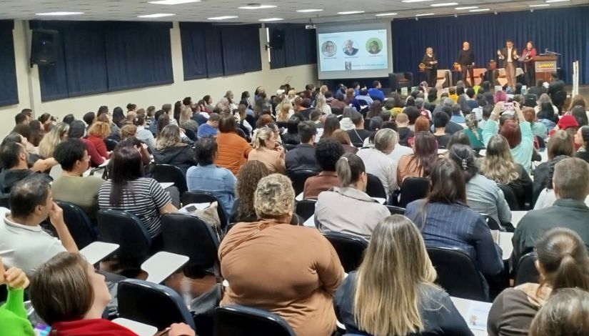 Fotografia de um auditório repleto de pessoas assistindo a uma palestra com 4 pessoas no palco e um telõa com 3 fotografias de pessoas do lado esquerdo.