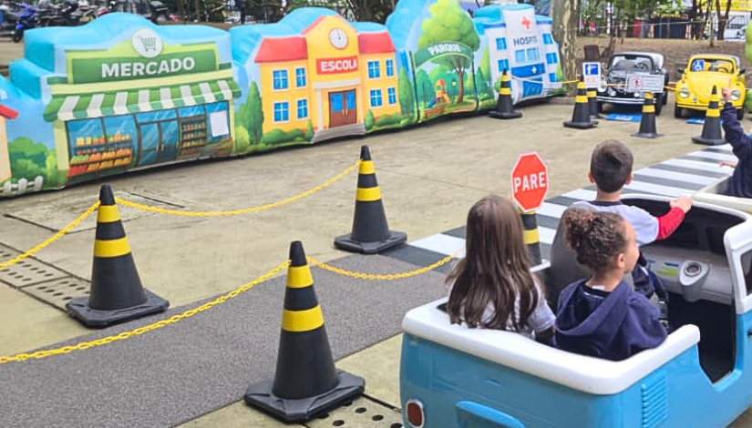 Fotografia de uma mini cidade com prédios, ruas, calçadas e sinalizações. Três crianças estão em um carrinho azul e passeiam pela cidade cenográfica.