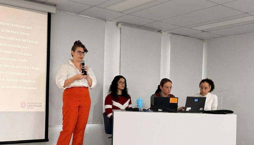 Fotografia de 4 mulheres ao palco. Uma está em pé e fala ao microfone e as outras três estão sentadas à mesa.