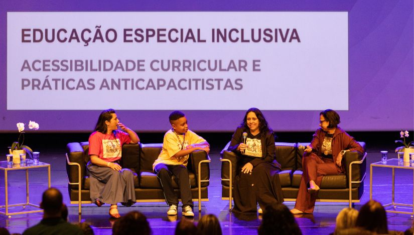 Fotografia do palco com quatro pessoas sentadas em dois sofás: são três mulheres e um menino. Ao fundo, o texto 