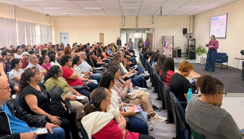 Fotografia de pessoas sentadas em um auditório, voltadas para uma mulher que está à frente do grupo e fala ao microfone.