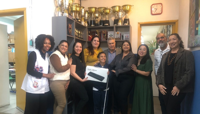 Grupo de pessoas reunidas em uma unidade escolar para a entrega de uma Linha Braille a uma estudante. Ao centro, a estudante segura uma caixa com a imagem do equipamento e uma bengala branca. Ao fundo, há uma estante com diversos troféus e medalhas.