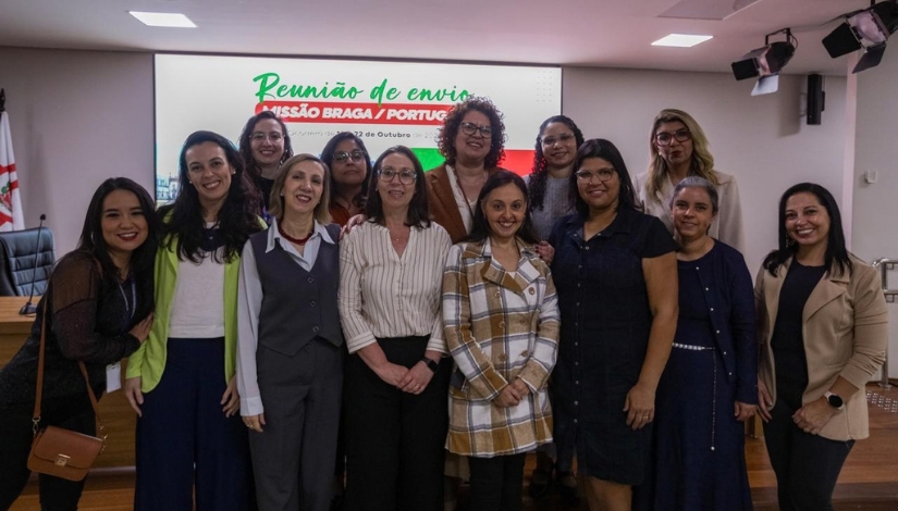 Grupo de mulheres reunidas em um auditório para registro fotográfico. Elas estão posicionadas em duas fileiras e sorriem para a câmera. Ao fundo, há uma tela de projeção com informações sobre a missão técnica entre São Paulo e Braga, em Portugal.