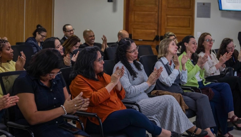Público acompanha uma atividade em um auditório. Pessoas sentadas em cadeiras aplaudem, enquanto algumas participantes sorriem e observam a apresentação. Ao fundo, há outras pessoas sentadas e uma parede de madeira.