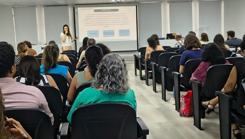 Auditório com diversas pessoas sentadas em cadeiras, acompanhando uma apresentação. Ao fundo, uma mulher fala ao microfone em frente a uma tela de projeção com informações sobre Ouvidoria. Ao lado da tela, há um computador e outros equipamentos utilizados durante a apresentação.