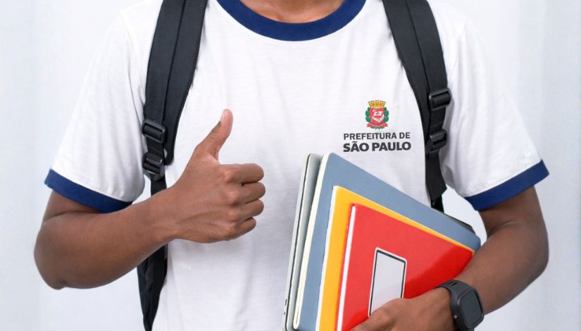Fotografia de um estudante fazendo sinal de joia com o polegar e segurando cadernos e livros. Ele usa uniforme da Prefeitura de São Paulo.