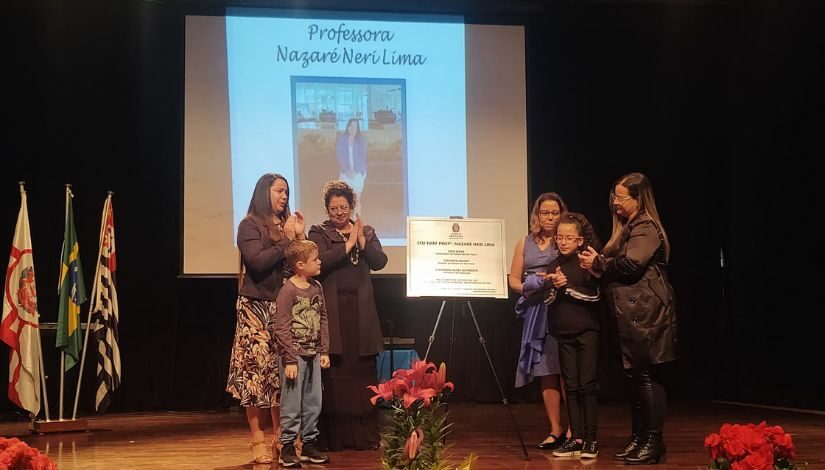 Fotografia do palco com seis pessoas, a placa de entronização da patronesse e ao fundo no telão projetado a imagem da Professora Nazaré Neri Lima.